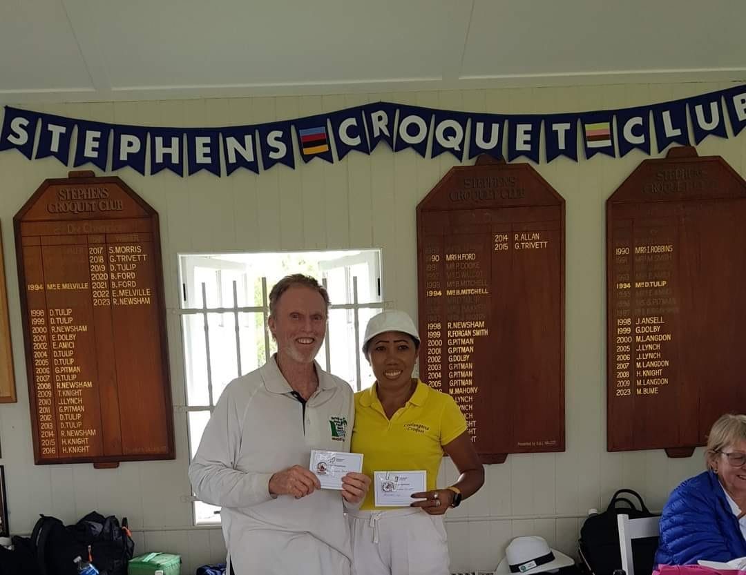 Two players hold prize cards in front of the Stephens Croquet Club honour boards, with winners' names going back to 1990.